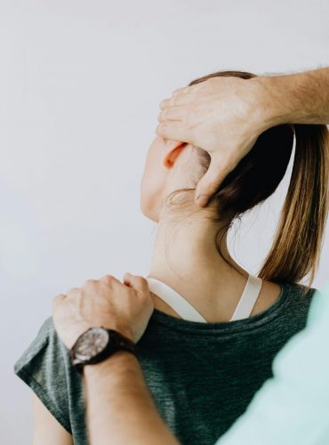 Back view of crop faceless chiropractor in wristwatch stretching neck of unrecognizable female patient during medical examination in doctor office in clinic